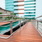 A imagem mostra uma passarela de madeira que leva a uma piscina. A passarela é feita de tábuas de madeira lisas e tem um corrimão de aço inoxidável. A piscina é retangular e tem uma borda de azulejos azuis. A água da piscina é azul clara e está limpa. Ao fundo, há um jardim com árvores e arbustos.