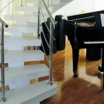 A imagem mostra uma escada branca com corrimão de aço inoxidável e um piano de cauda em uma sala de estar