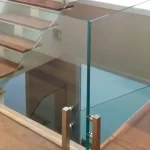 A imagem mostra uma escada com degraus de madeira e guarda-corpos de vidro. Os painéis de vidro são fixados por suportes de metal inoxidável, que estão presos ao chão de madeira. A combinação de madeira e vidro, junto com os detalhes em inox, cria um visual moderno e elegante. A escada leva a um nível inferior, e o guarda-corpo de vidro proporciona segurança enquanto mantém uma aparência leve e transparente.