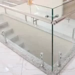 A imagem mostra uma escada com um guarda-corpo de vidro e corrimão de aço inoxidável. A escada é cercada por painéis de vidro fixados por suportes de metal inoxidável, que estão presos a uma base de granito. O corrimão de aço inoxidável está conectado aos painéis de vidro, proporcionando segurança e um visual elegante e moderno.